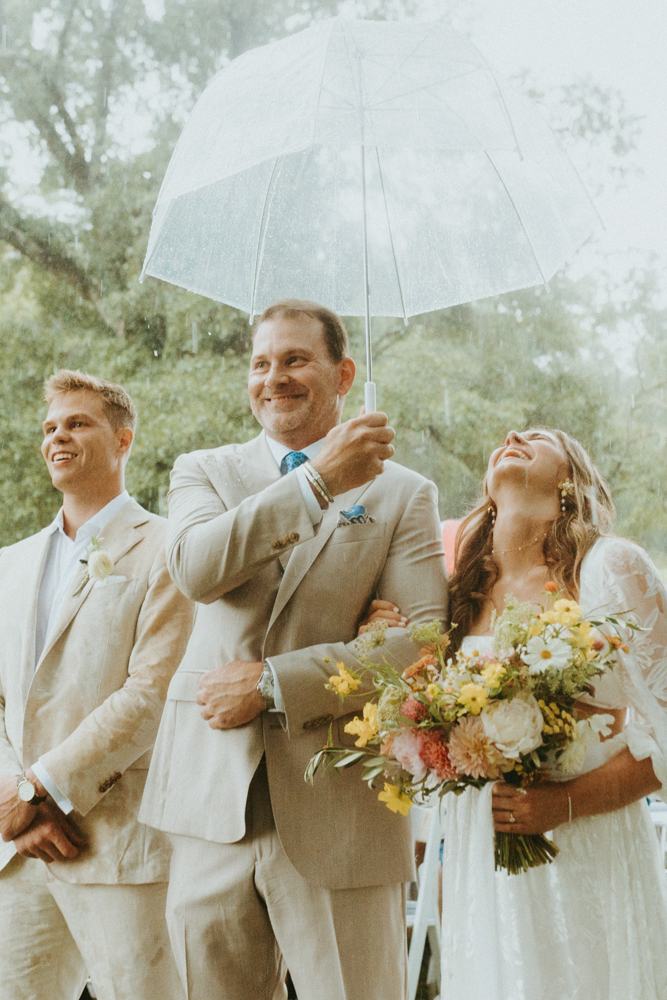 The father of the bride holds an umbrella over the bride as he gives her away at the wedding ceremony.