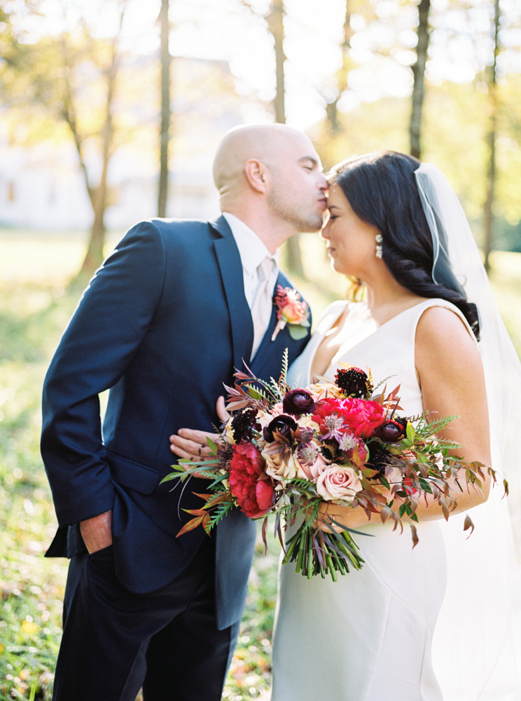 The groom kisses the bride's forehead as she holds her fall bouquet of flowers for their wedding in Birmingham.