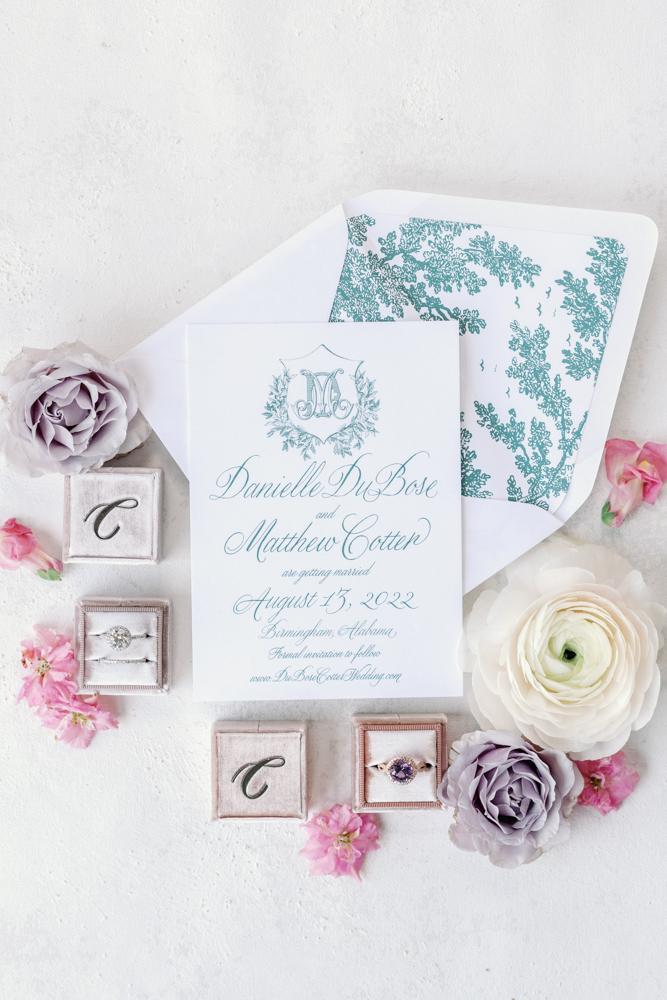 The wedding invitation is photographed in a lay flat style with the wedding rings and flowers.