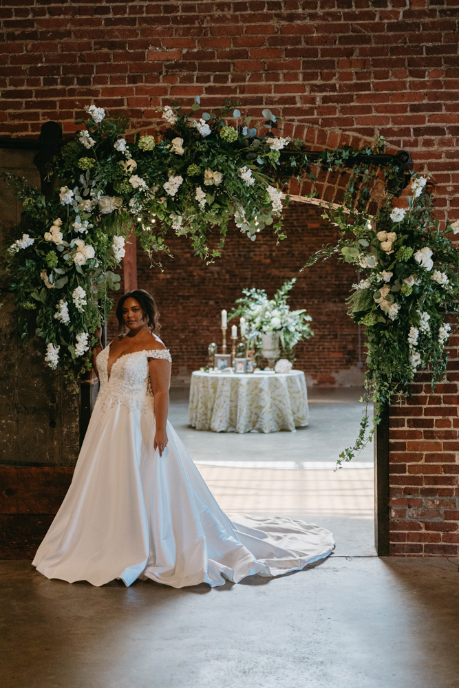 The bride stands beneath the floral arch at B&A Warehouse.