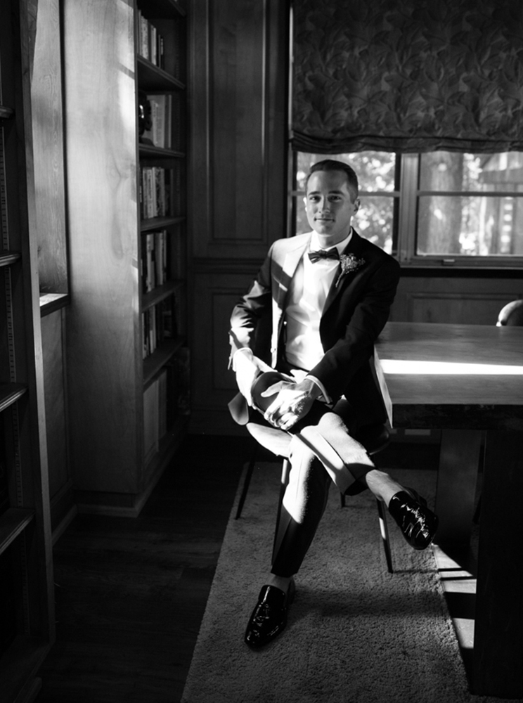 The groom sits by the window in his tuxedo.