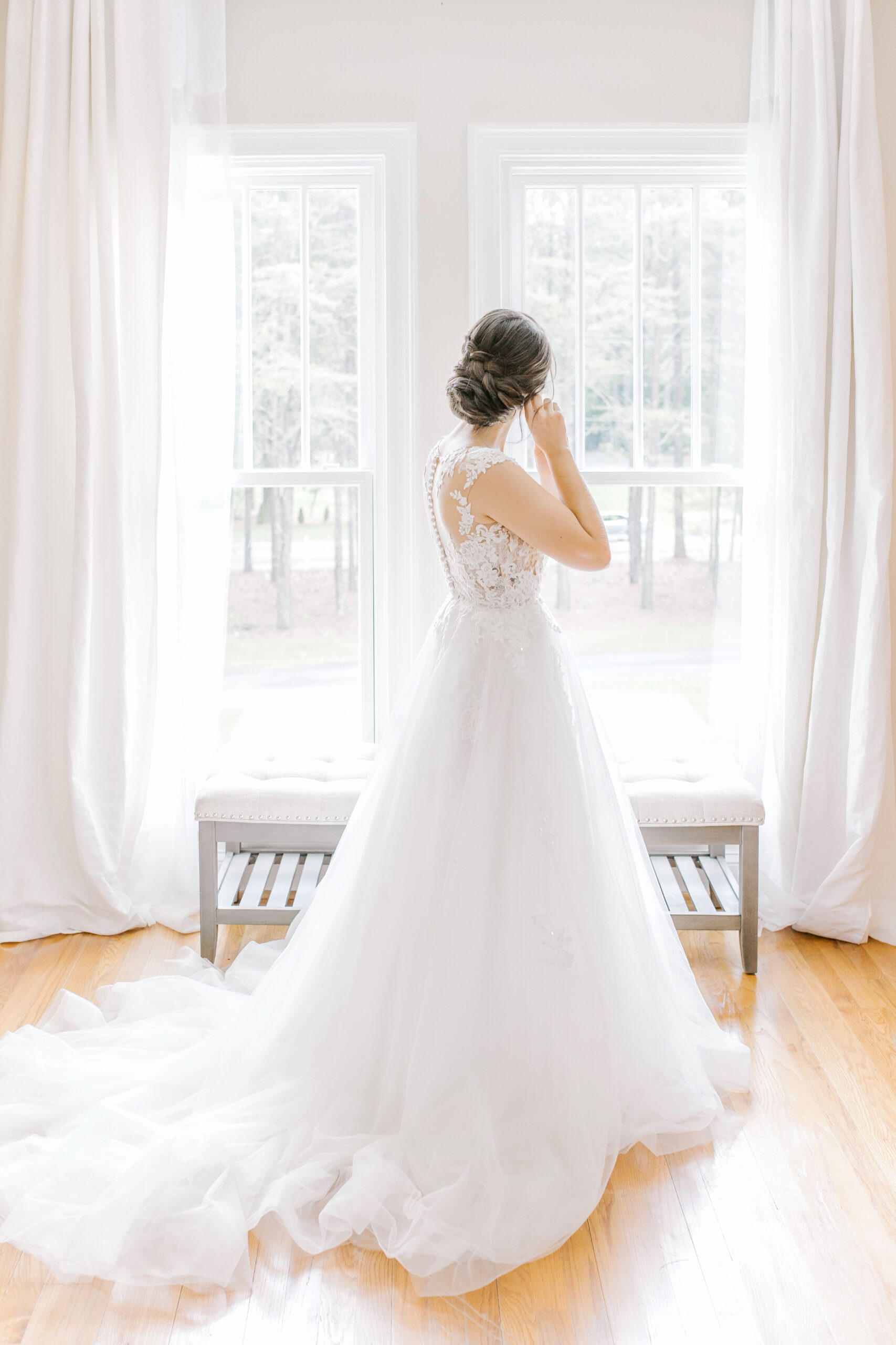 The bride puts on her earrings while looking out the window.