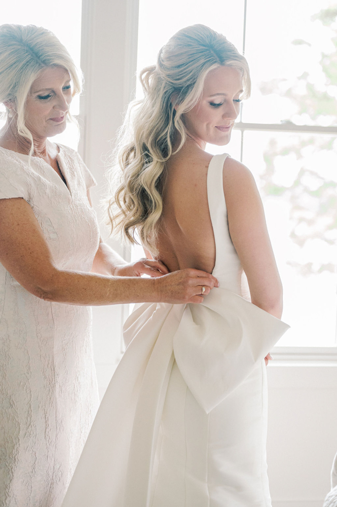 The mother of the bride helps her daughter into her wedding dress with a large bow on the back.